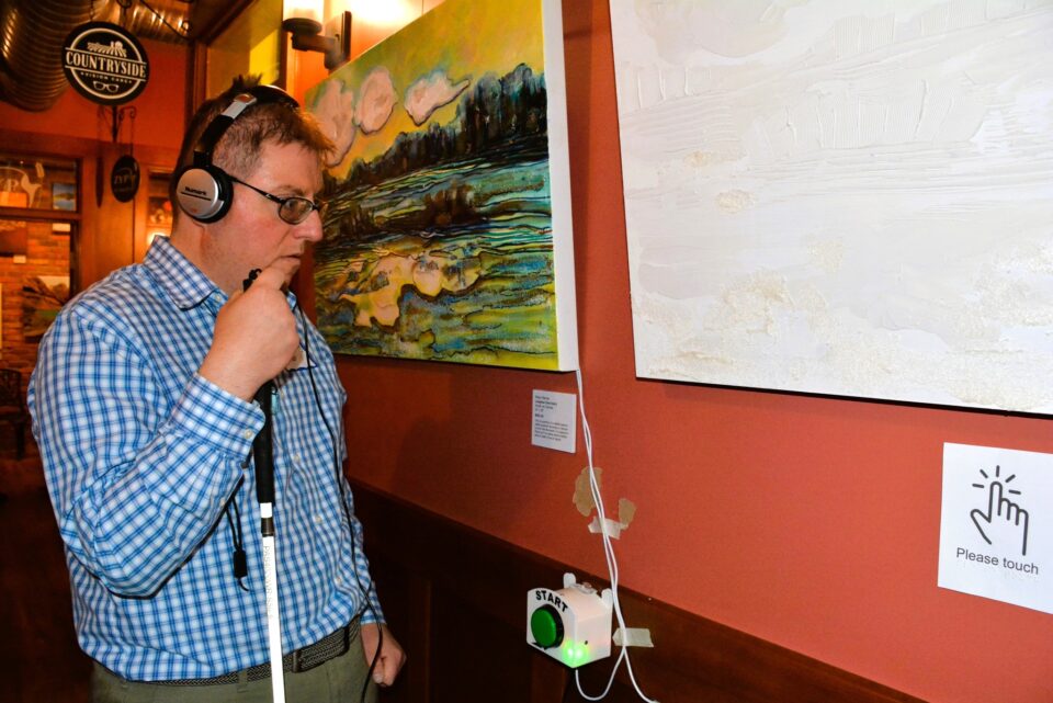 A man holding a mobility cane stands beside a tactile replica while listening to an audio description